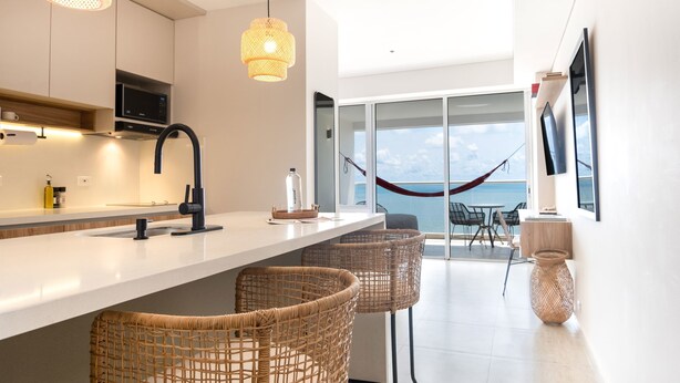 Cocina moderna con encimera blanca, grifo negro y sillas de mimbre. Una ventana grande ofrece vistas al mar y hay una hamaca.