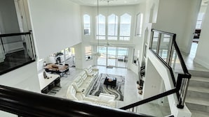 A spacious living area with a high ceiling, a staircase, and large windows overlooking a body of water.