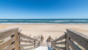 Ein Holzsteg führt zu einem Sandstrand, im Hintergrund ist das Meer zu sehen.