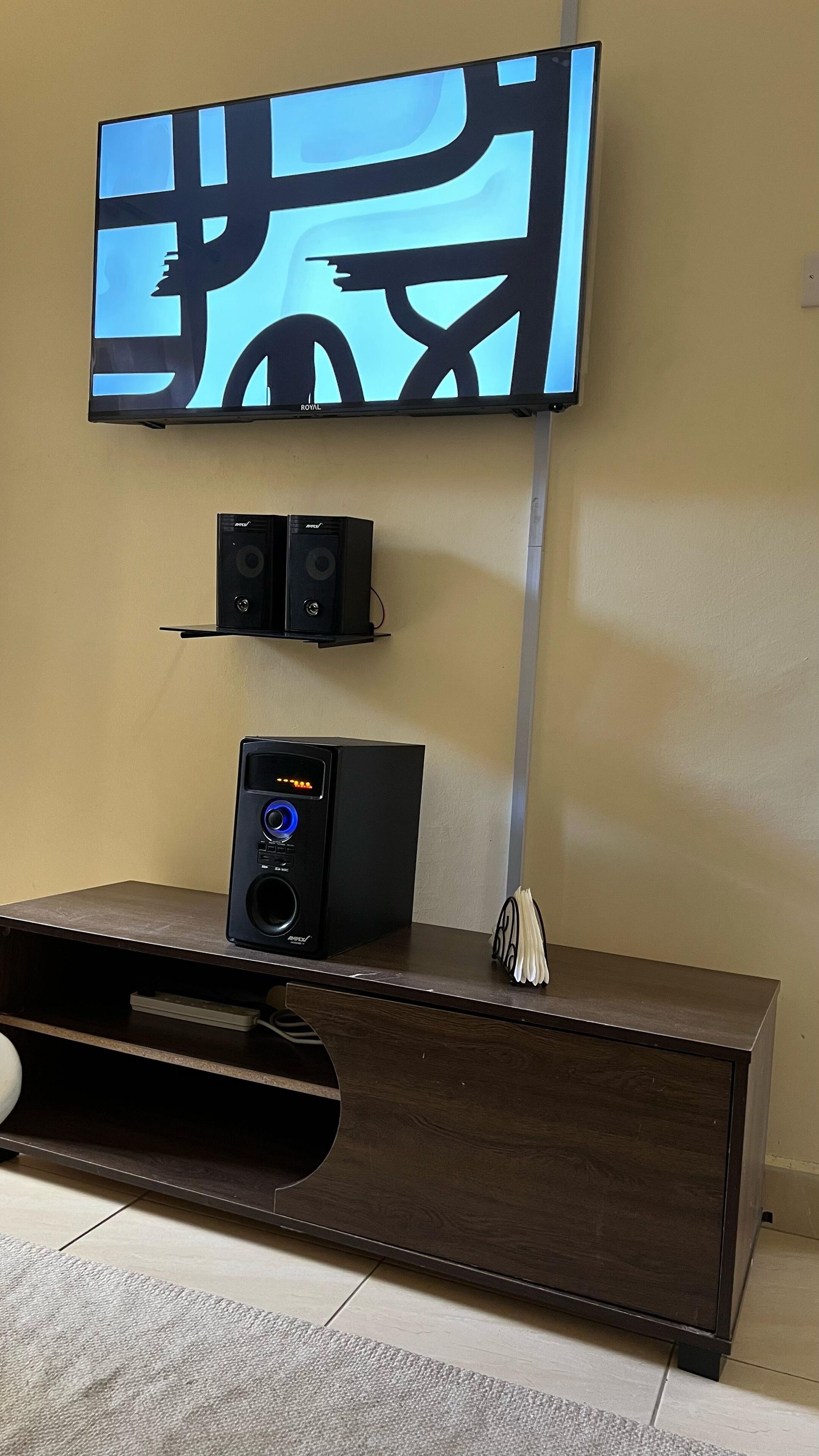 A home theater setup with a flat-screen TV mounted on the wall, two speakers on a shelf below, and a subwoofer on a wooden cabinet.