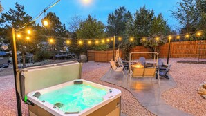 Un bain à remous avec une terrasse en bois, entouré d’une clôture en bois, de guirlandes lumineuses et d’un espace patio avec des chaises et un foyer.