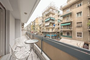 Un balcon avec des chaises blanches et une petite table, d’où l’on a vue sur une rangée d’immeubles à plusieurs étages, dotés de balcons et d’auvents.