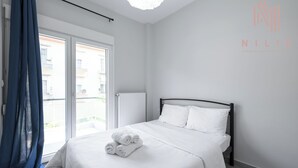Une chambre d’hôtel avec un lit, du linge de lit blanc, une fenêtre donnant sur des immeubles et un rideau bleu.