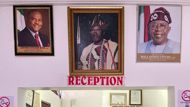 A reception area with framed portraits, a 'RECEPTION' sign, a no smoking sign, and a reception desk with two staff members. - Abuja Bays Garden Hotel (Abuja)