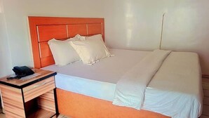 A single bed with a wooden headboard, a bedside table with a phone, and a plain wall. - Abuja Bays Garden Hotel (Abuja)