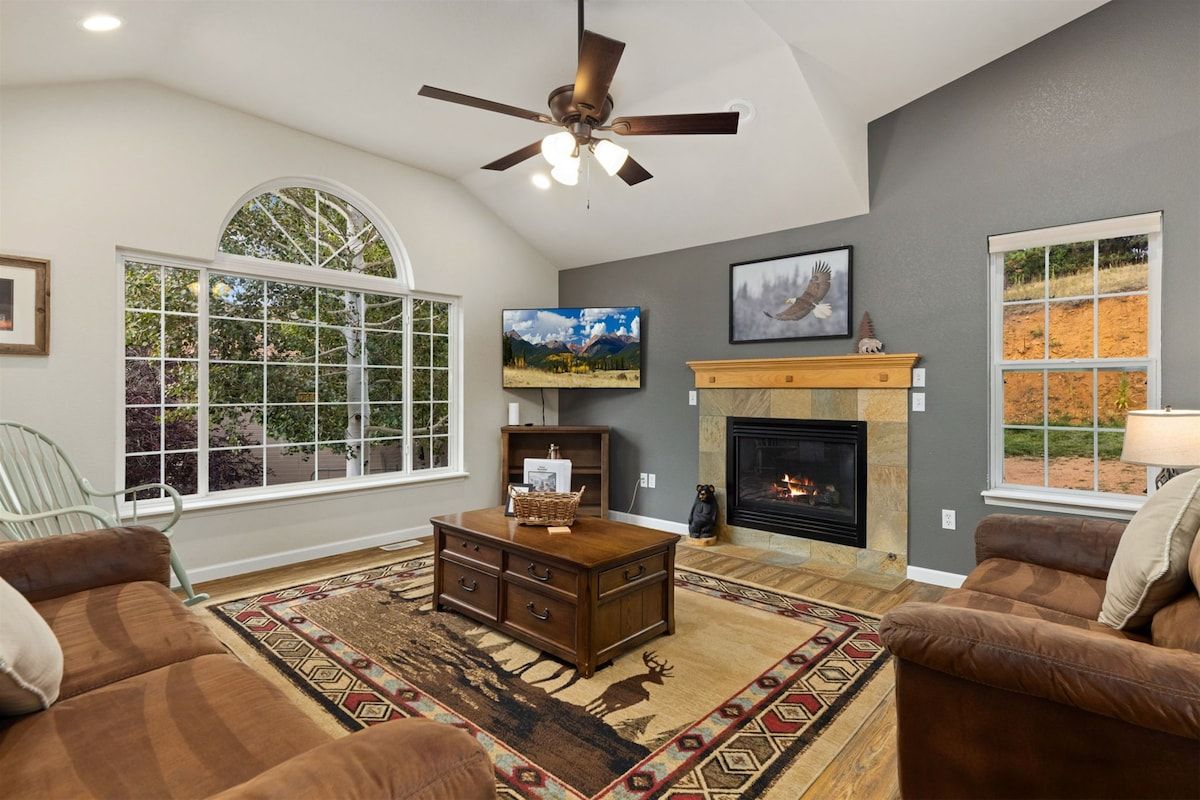 A living room with a fireplace, a ceiling fan, and a large window.