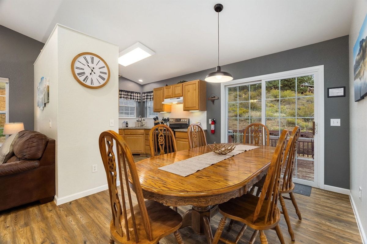 A dining area with a wooden table and chairs, a clock on the wall, and a kitchen in the background.