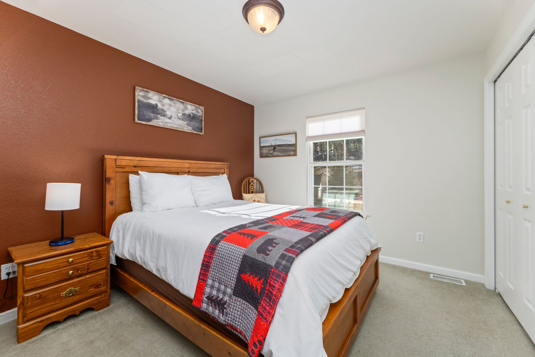 A bedroom with a wooden bed, white bedding, a red and black quilt, a nightstand with a lamp, and a window with a view of trees.