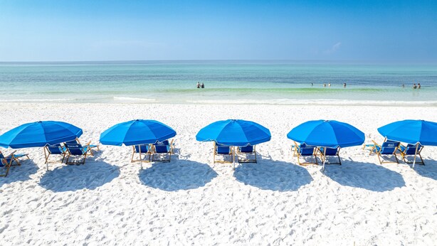 A beach with white sand and a clear blue sky. There are five blue umbrellas and matching lounge chairs arranged in a row.