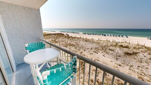 A balcony with a white table and turquoise chairs, overlooking a beach with umbrellas and people.