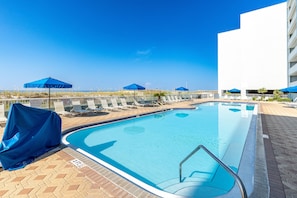 Una piscina con lettini e ombrelloni, cielo sereno e un edificio sullo sfondo.