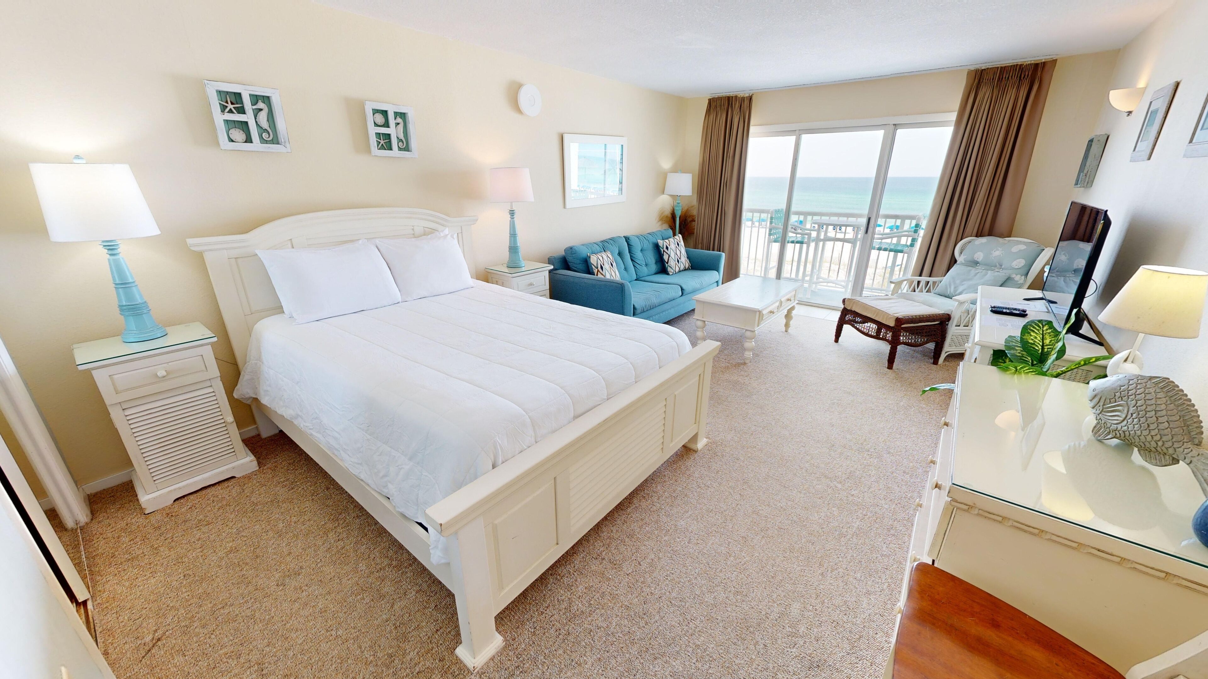 A hotel room with a large bed, a sofa, a desk with a laptop, and a view of the ocean.