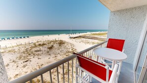 A balcony with a white table and red chairs overlooking a beach with blue umbrellas and a wooden walkway. - Islander Beach Resort 302 Condo by RedAwning (Fort Walton Beach)