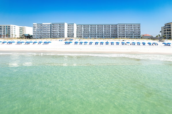 Un hotel frente a la playa con varios pisos, una playa de arena y sombrillas azules.