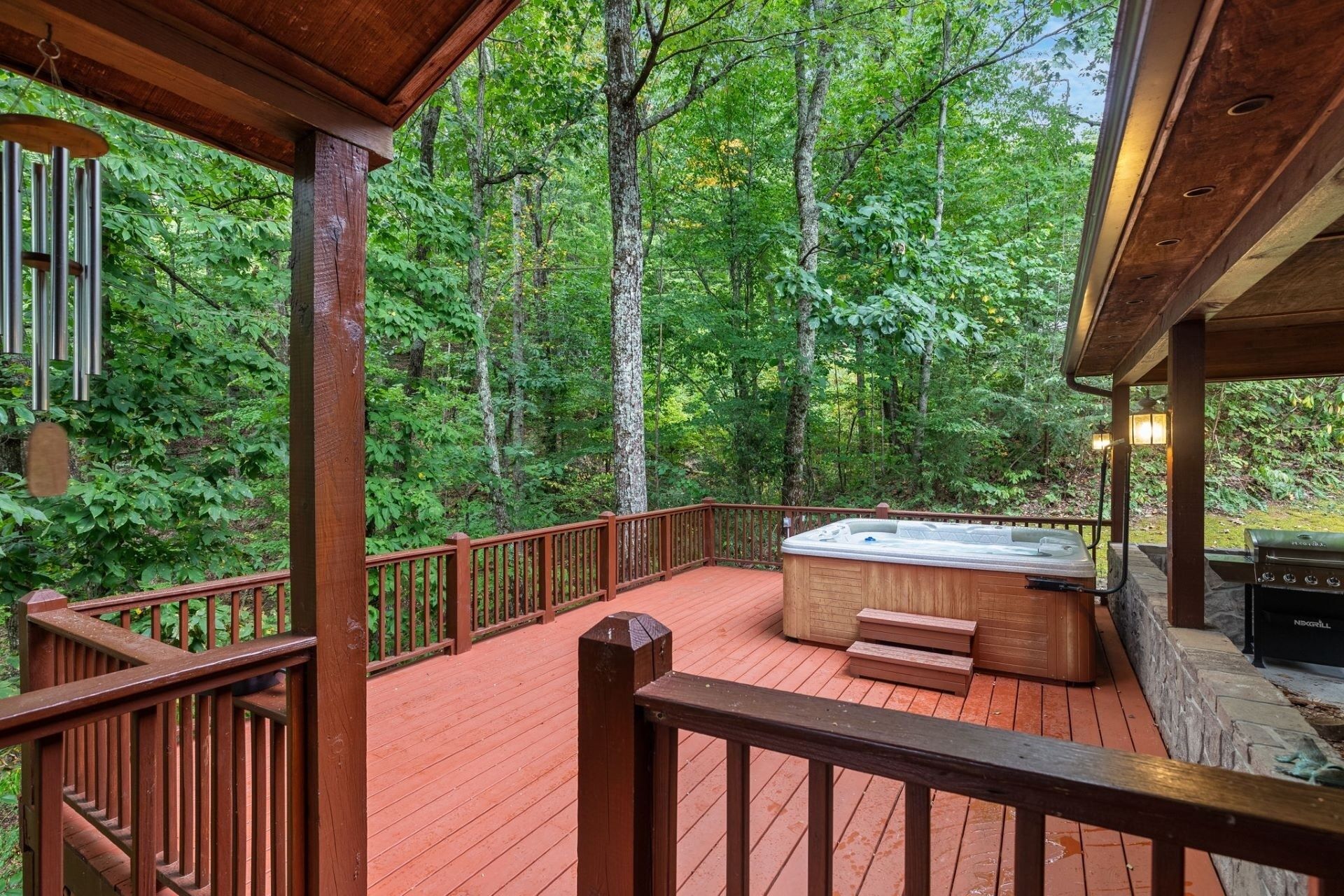 A wooden deck with a hot tub and barbecue grill, surrounded by a forested area.