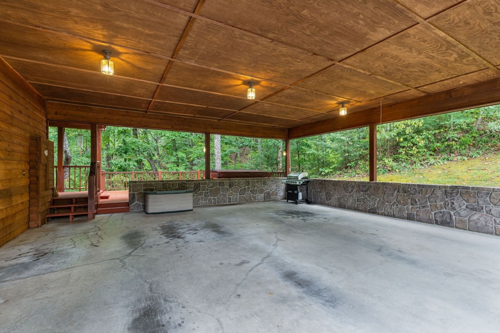 A covered outdoor area with a stone wall, a grill, and a concrete floor.