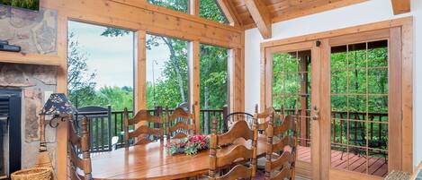 A wooden dining table with chairs, a stone fireplace, and large windows offering a view of the surrounding forest.