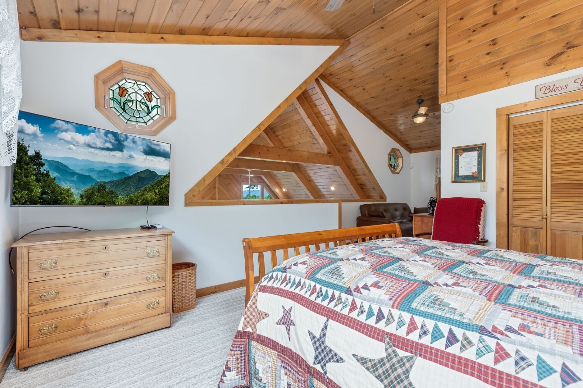 Un dormitorio con techo de madera, una cama con colcha colorida, un aparador, un televisor y una ventana con vista a la montaña.