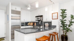 A modern kitchen with white cabinets, a stainless steel refrigerator, a microwave, and a granite countertop. There are two wooden stools and a potted plant. - Big Island Ali'i Villas 333 2 Bedroom Condo (Kailua-Kona)