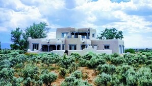 Una casa moderna de dos pisos con techo plano y varios balcones, rodeada de arbustos y árboles frondosos.