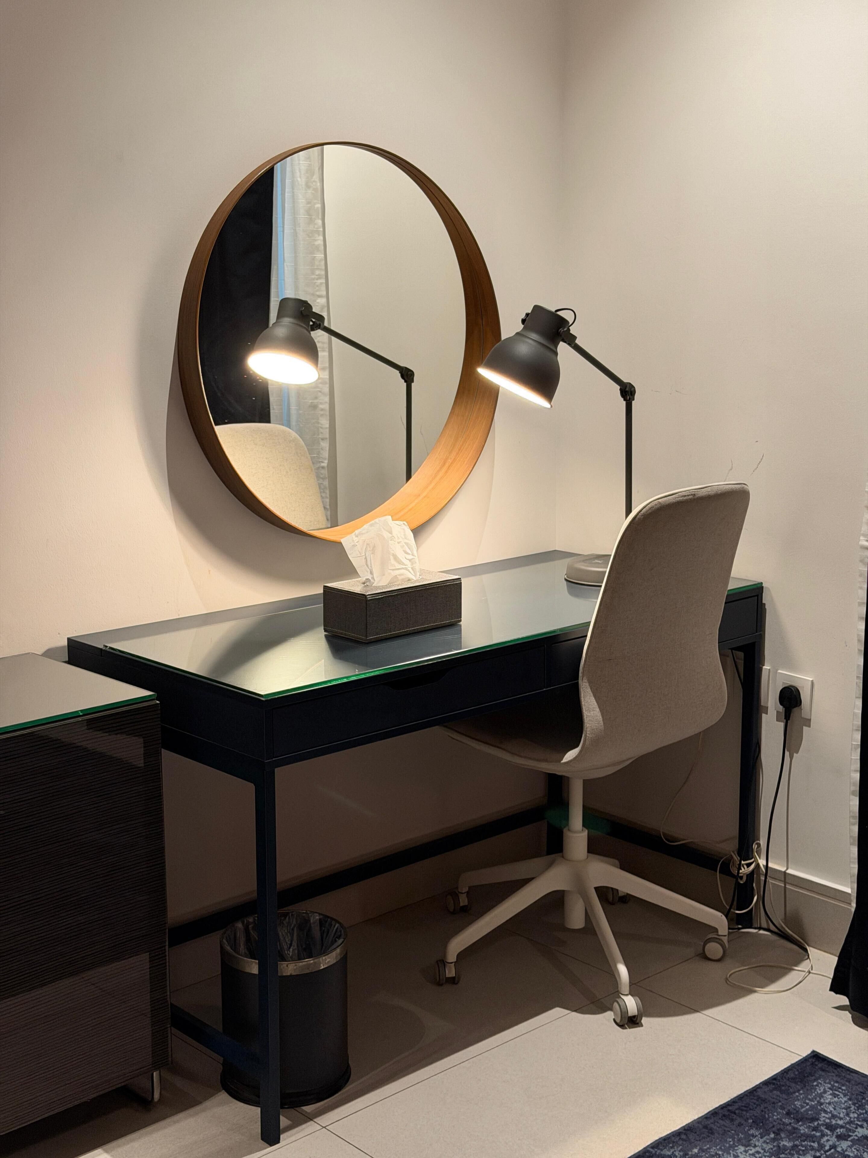A modern office desk with a round mirror, two desk lamps, a chair, and a trash can.