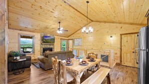 A wooden cabin interior with a dining table set for four, a sofa, a fireplace, and a kitchen area. - 4/4 Cabin w 4 King Suites|Hot Tub|Views 5 min to Gatlinburg! (Gatlinburg)