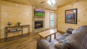 A cozy cabin interior with a stone fireplace, wooden walls, a leather couch, and a television mounted on the wall. - 4/4 Cabin w 4 King Suites|Hot Tub|Views 5 min to Gatlinburg! (Gatlinburg)