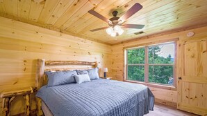 A wooden cabin bedroom with a bed, a ceiling fan, and a window with a view of trees. - 4/4 Cabin w 4 King Suites|Hot Tub|Views 5 min to Gatlinburg! (Gatlinburg)