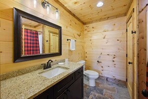 Baño de cabaña de madera con lavabo de granito, espejo, inodoro y cortina de ducha.