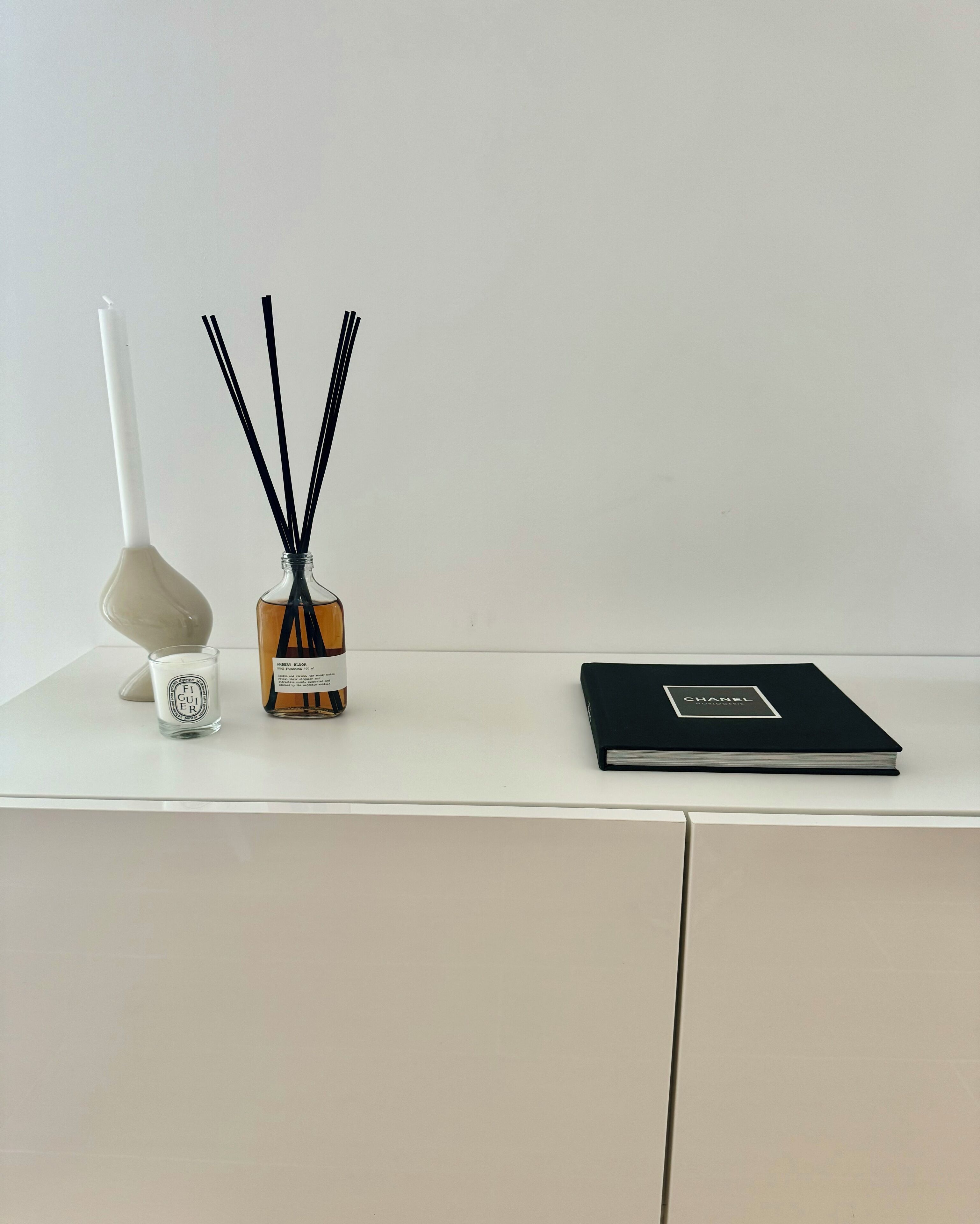Un bureau blanc orné d’une bougie blanche, d’un diffuseur et d’un livre noir portant le logo Chanel.