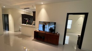 A modern living room with a flat-screen TV on a wooden stand, a kitchen area with a hanging light, and a hallway leading to a door. - Amore Villa Retreat in Ocho Rios (Ocho Rios)