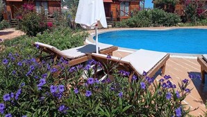 A poolside area with lounge chairs and an umbrella, surrounded by flowers and wooden buildings with red roofs. - Habesa Bungalov & Apart (Kas)