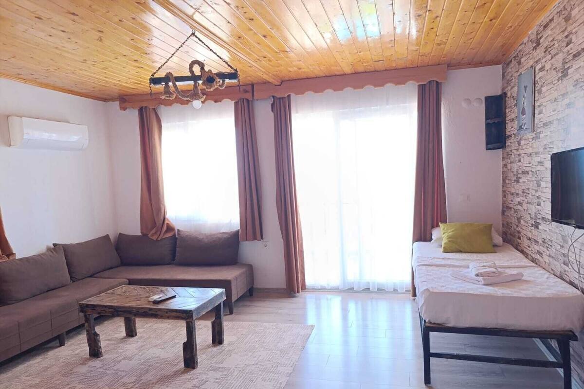 A cozy living room with a wooden ceiling, a sofa, a coffee table, and a bed with a TV on the wall.
