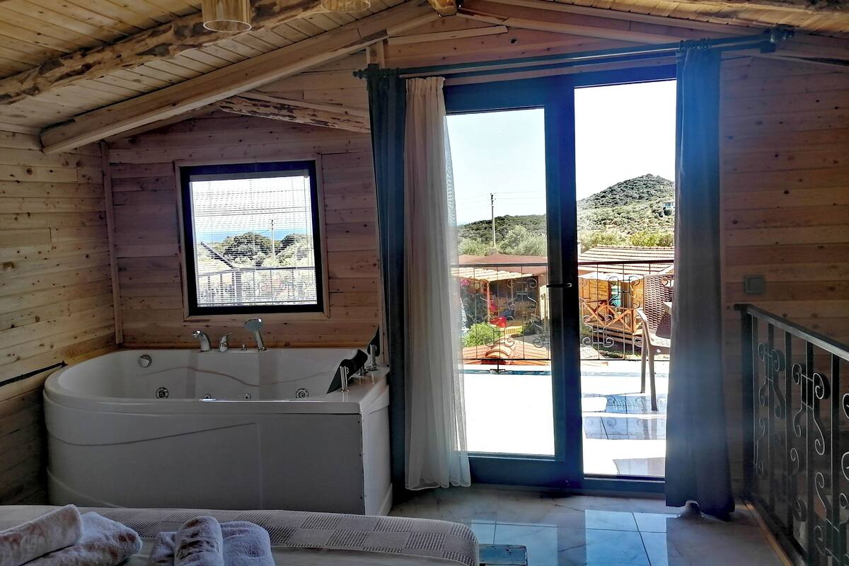 A wooden cabin with a large bathtub, a window with a view of the outside, and a balcony with a railing.