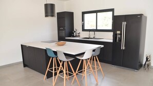 A modern kitchen with a central island, black cabinetry, a window, and a refrigerator. - Villa Venturon Proche la Plage de Pinarello Avec Piscine et Cuisine D'été (Zonza)