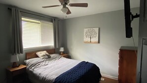 Een slaapkamer met een bed, een plafondventilator, een nachtkastje met lamp, een raam met blinds, een aan de muur bevestigde televisie en een houten kast.
