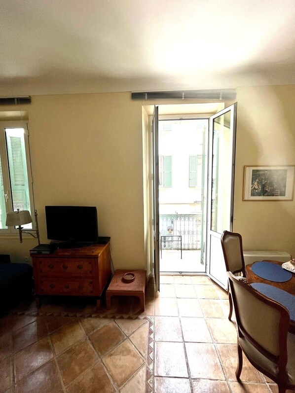 A room with a television on a wooden cabinet, a small table with a chair, and a balcony with a view of buildings. - Aufenthalt im Herzen von Cannes (Cannes)
