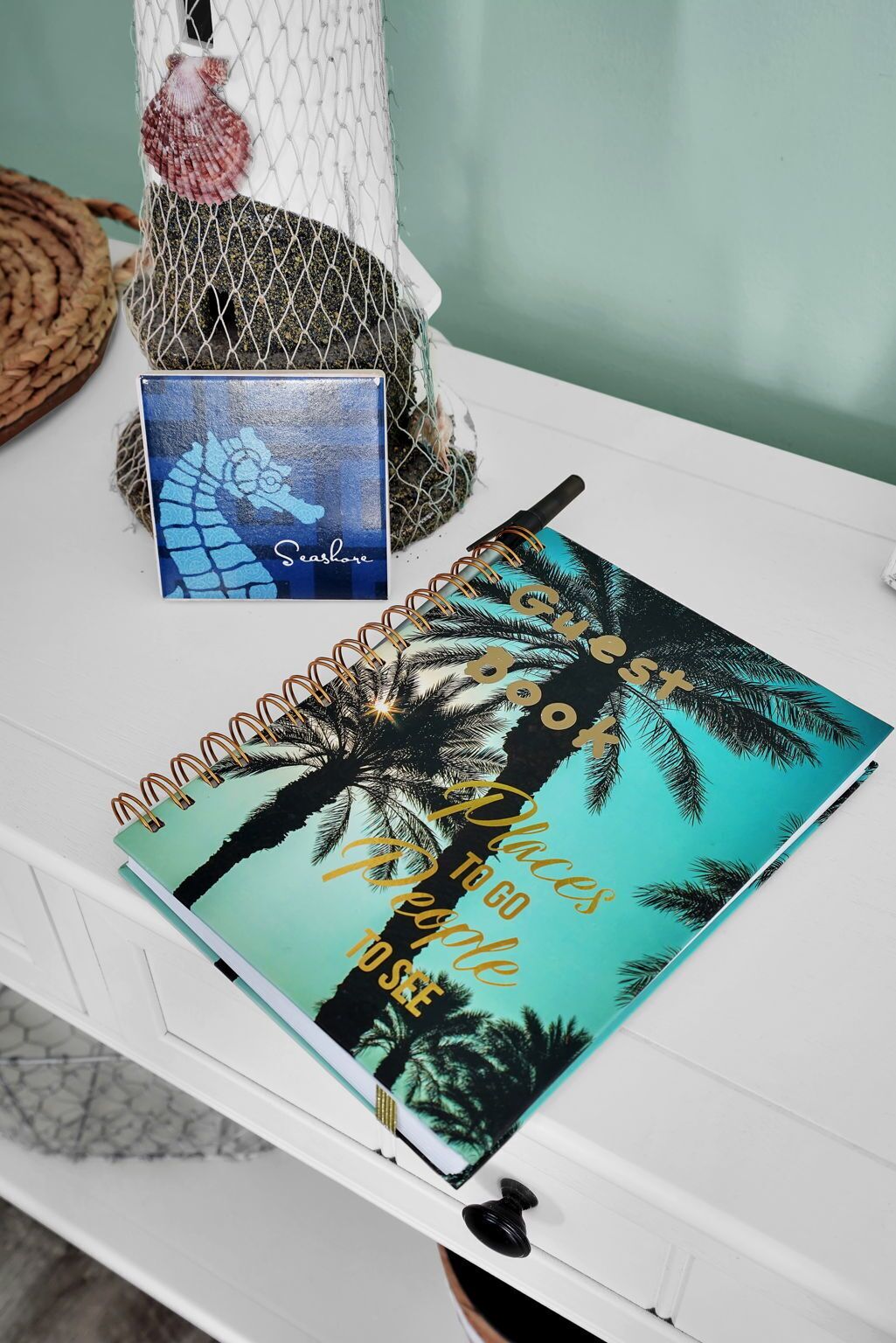 Un carnet avec un motif représentant un palmier tropical et l’inscription « Lieux à visiter, personnes à rencontrer » posé sur une table blanche.
