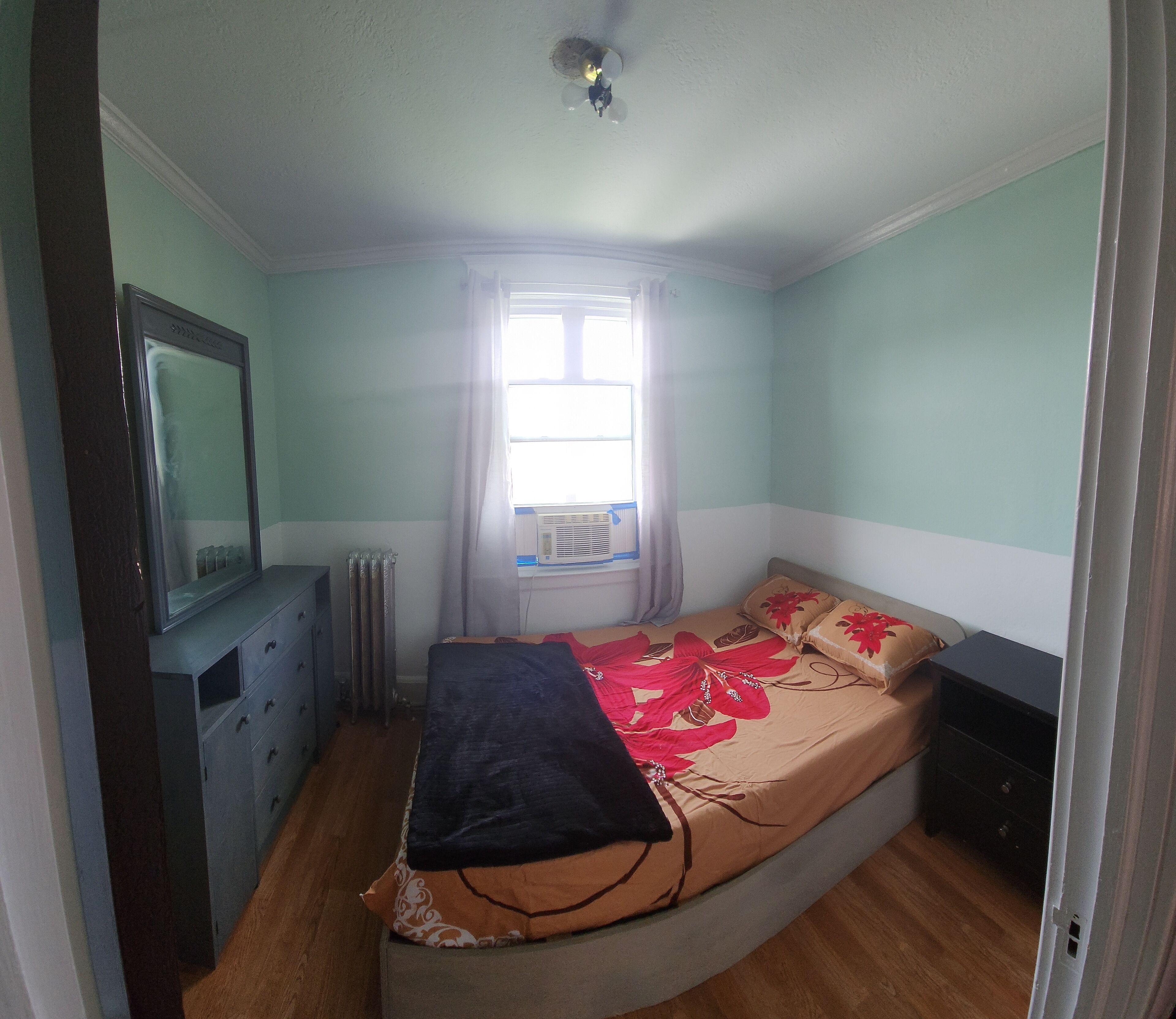 A bedroom with a bed, a dresser, a mirror, a radiator, and a window with curtains.
