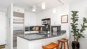 A modern kitchen with white cabinets, a stainless steel refrigerator, a microwave, and a granite countertop. There are two wooden stools and a potted plant. - Oceanfront Kona Condo | Watch Whales & Sunsets from Your Private Lanai (Kailua-Kona)