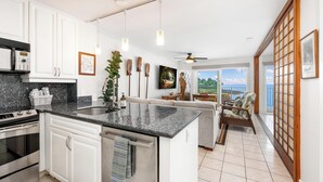 A modern kitchen with white cabinets, a granite countertop, and stainless steel appliances. There is a dining area with a view of the outdoors. - Oceanfront Kona Condo | Watch Whales & Sunsets from Your Private Lanai (Kailua-Kona)