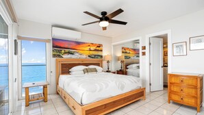 A bedroom with a bed, a ceiling fan, a nightstand, a wooden dresser, a small table, and a large window with a view of the ocean. - Oceanfront Kona Condo | Watch Whales & Sunsets from Your Private Lanai (Kailua-Kona)