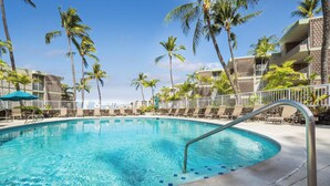 A swimming pool with a metal handrail, surrounded by lounge chairs and palm trees, with a building in the background. - Oceanfront Kona Condo | Watch Whales & Sunsets from Your Private Lanai (Kailua-Kona)