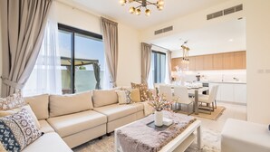 A modern living room with a beige sofa, a coffee table with a vase, and a dining area with a white table and chairs.