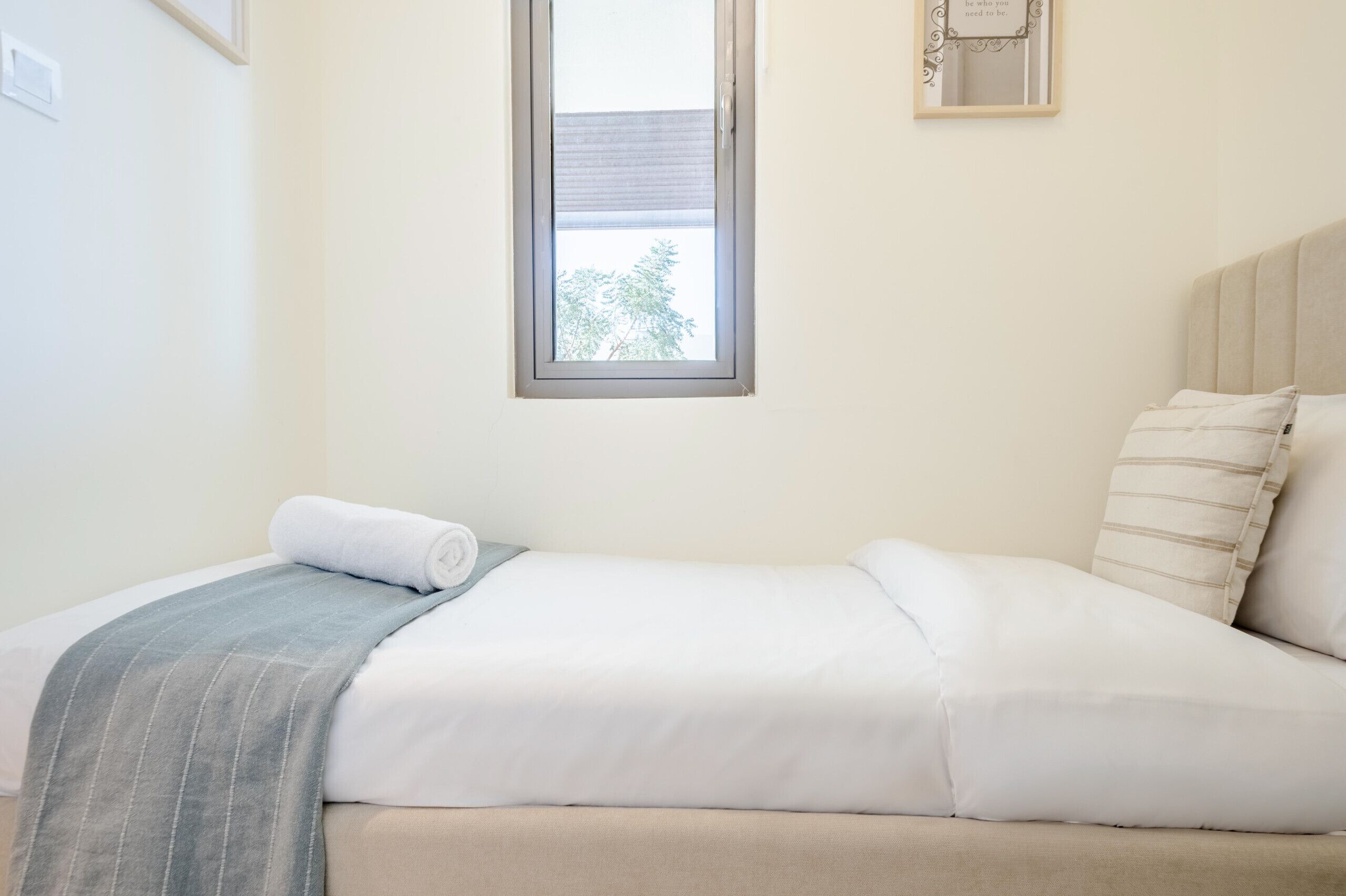 A neatly made bed with a rolled towel and a gray blanket, a window with a view of trees, and a framed quote on the wall.