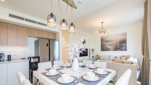 A modern kitchen with built-in appliances, a dining area with a white table set for six, and a living area with a sofa and a large landscape painting.
