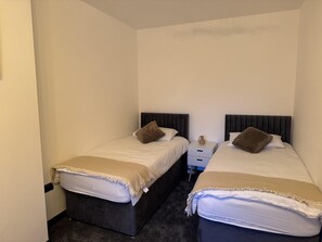 Une petite chambre propre, avec deux lits, chacun doté d’une tête de lit sombre et d’une couverture beige. Une petite table se trouve entre les lits, et un vase y est posé.