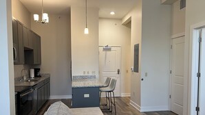 A modern kitchen with granite countertops, a built-in oven, and a coffee maker. There is a gray sofa and a small island with bar stools. The room has wooden flooring and a white door. - Downtown Adrian apartment with parking and wifi (Adrian)