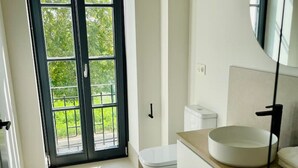 A modern bathroom with a toilet, sink, and mirror. There is a large window with a view of greenery outside. - Sueño Indiano, Bajo Norte (Muros de Nalón)
