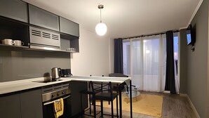 Cuina compacta amb una illa central, zona de menjador amb dues cadires, un televisor fixat a la paret i una finestra gran amb cortines.